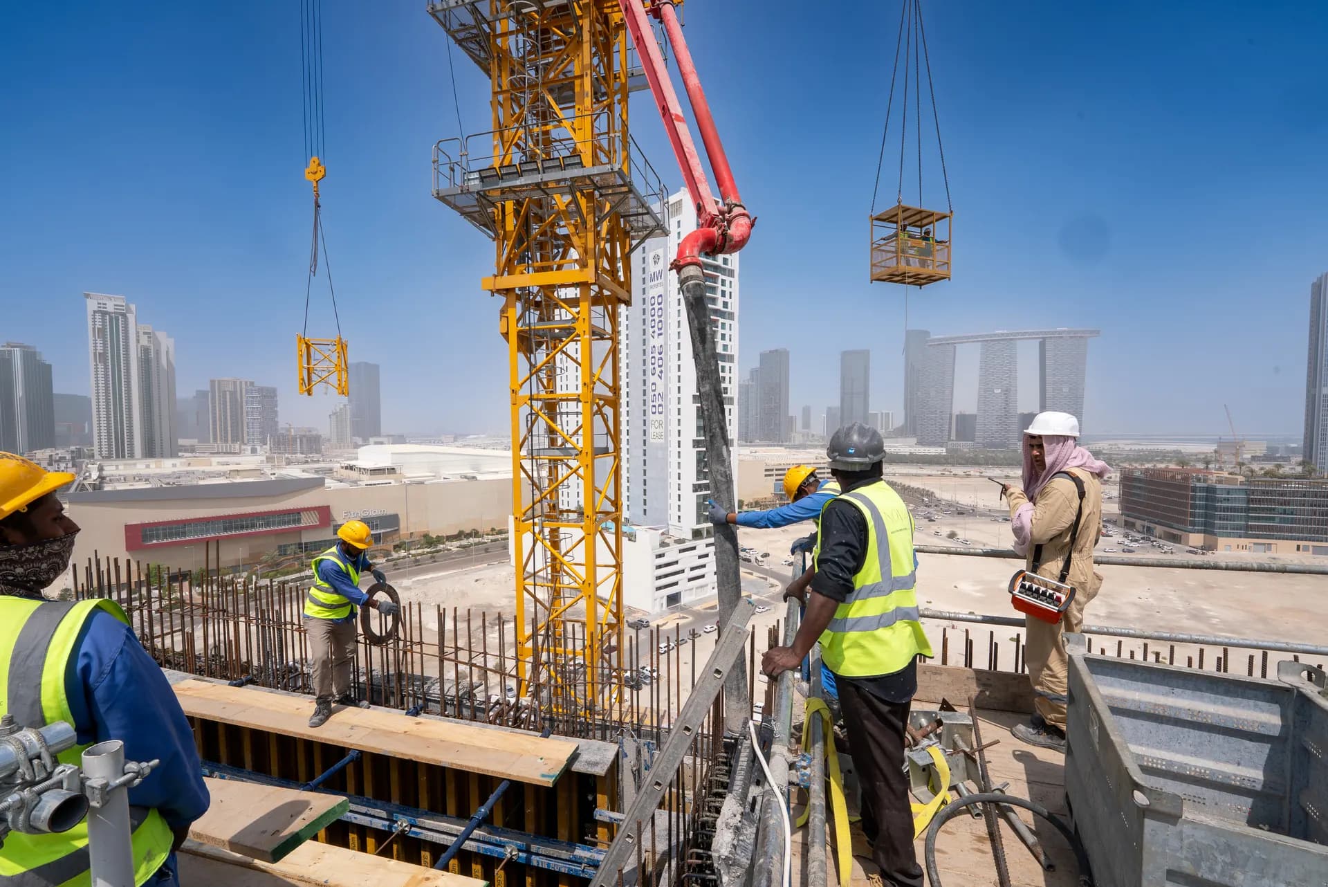 Construction crew pouring concrete and tying rebar on a high-rise building site, with a tower crane and the Abu Dhabi skyline in the background